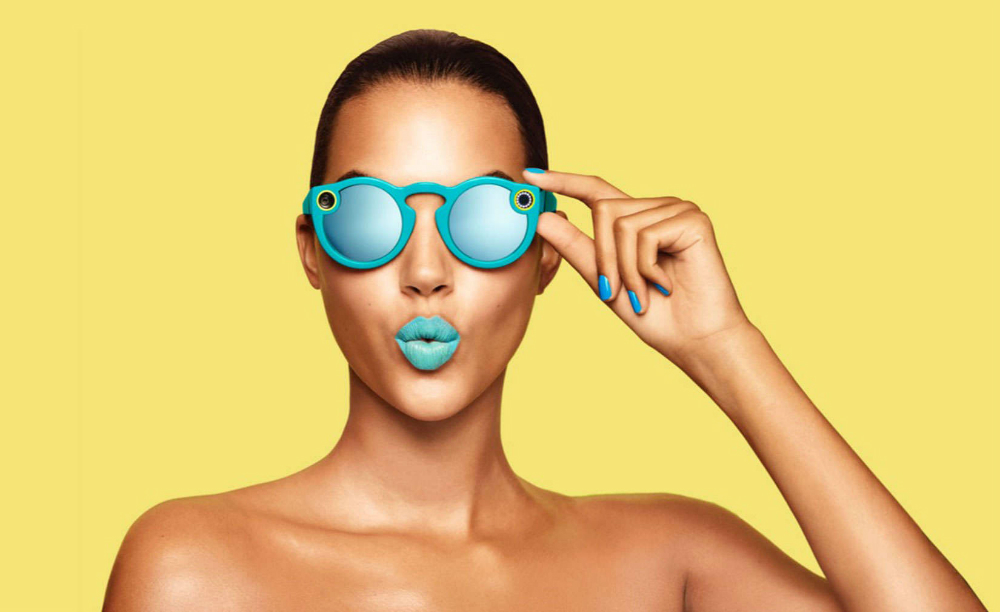 Snapchat's video recording glasses. A woman is wearing the bright blue glasses, which matching bright blue lipstick and nails against a yellow background.